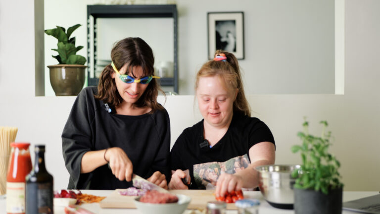 Mona Arzberger und Hanna Gugler stehen nebeinander in einer Küche. Mona trägt eine Taucherbrille und schneidet Zwiebel. Hanna steht neben ihr und greift nach Tomaten. Auf der Küchenoberfläche stehen Tomatemsoße, Gemüse und Spagetti.